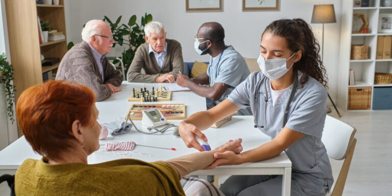 Female Nurse Checking Blood Pressure of Elderly Woman in a Shared Activity Room at an Assisted Living Facility